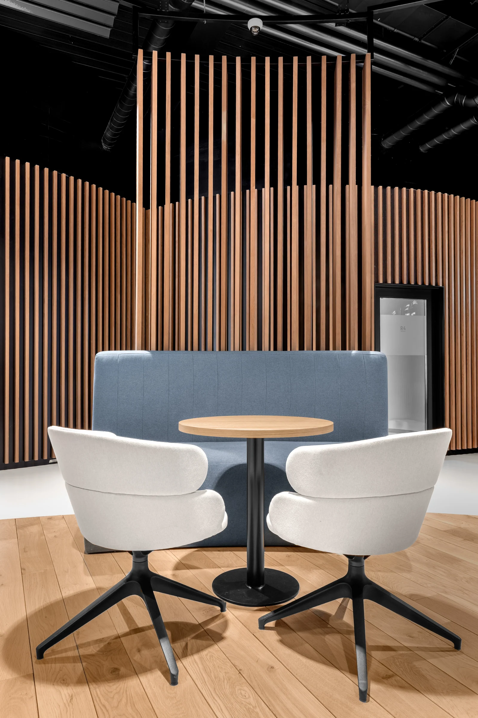 Une table ronde, deux fauteuils blancs et une banquette à dossier bleu dans une alcôve habillée de lames verticales.