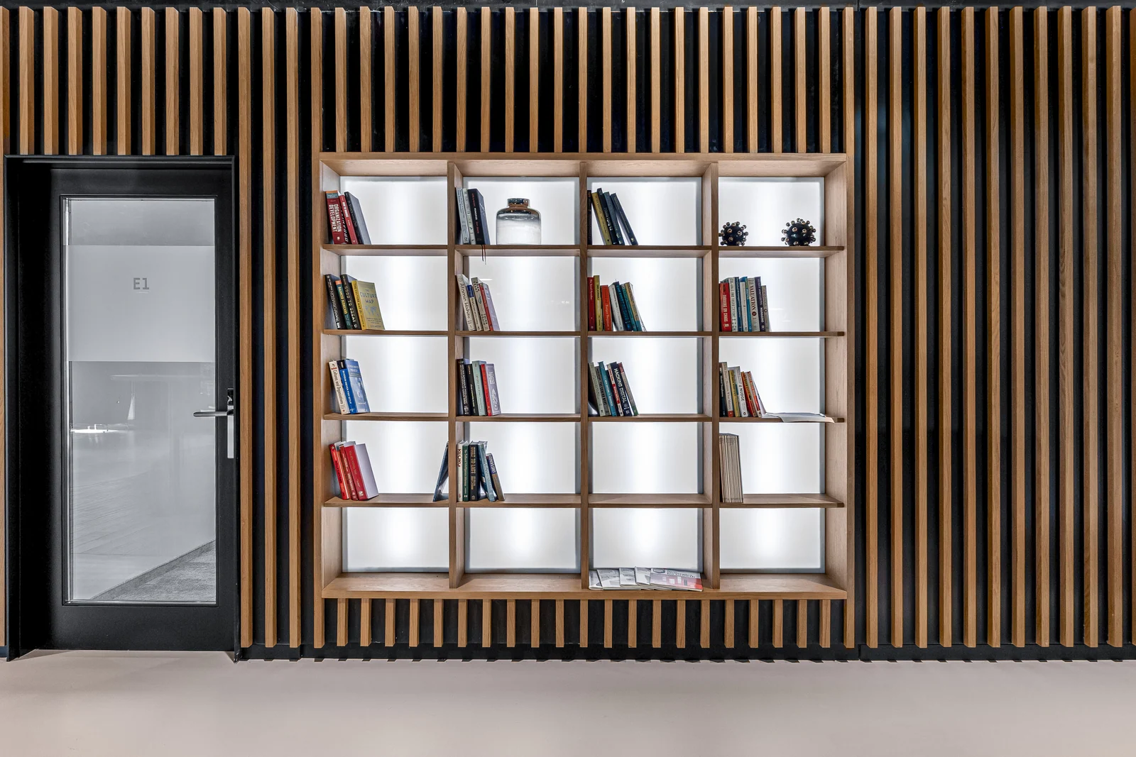 Bibliothèque à cases ouvertes intégrée dans une paroi en lames de bois, à côté d’une porte vitrée.