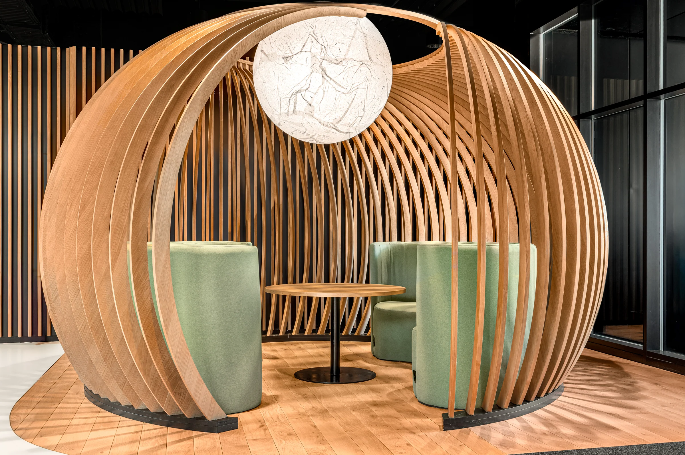 Des lames de bois dessinent une alcôve arrondie autour d’une table et de deux fauteuils verts.